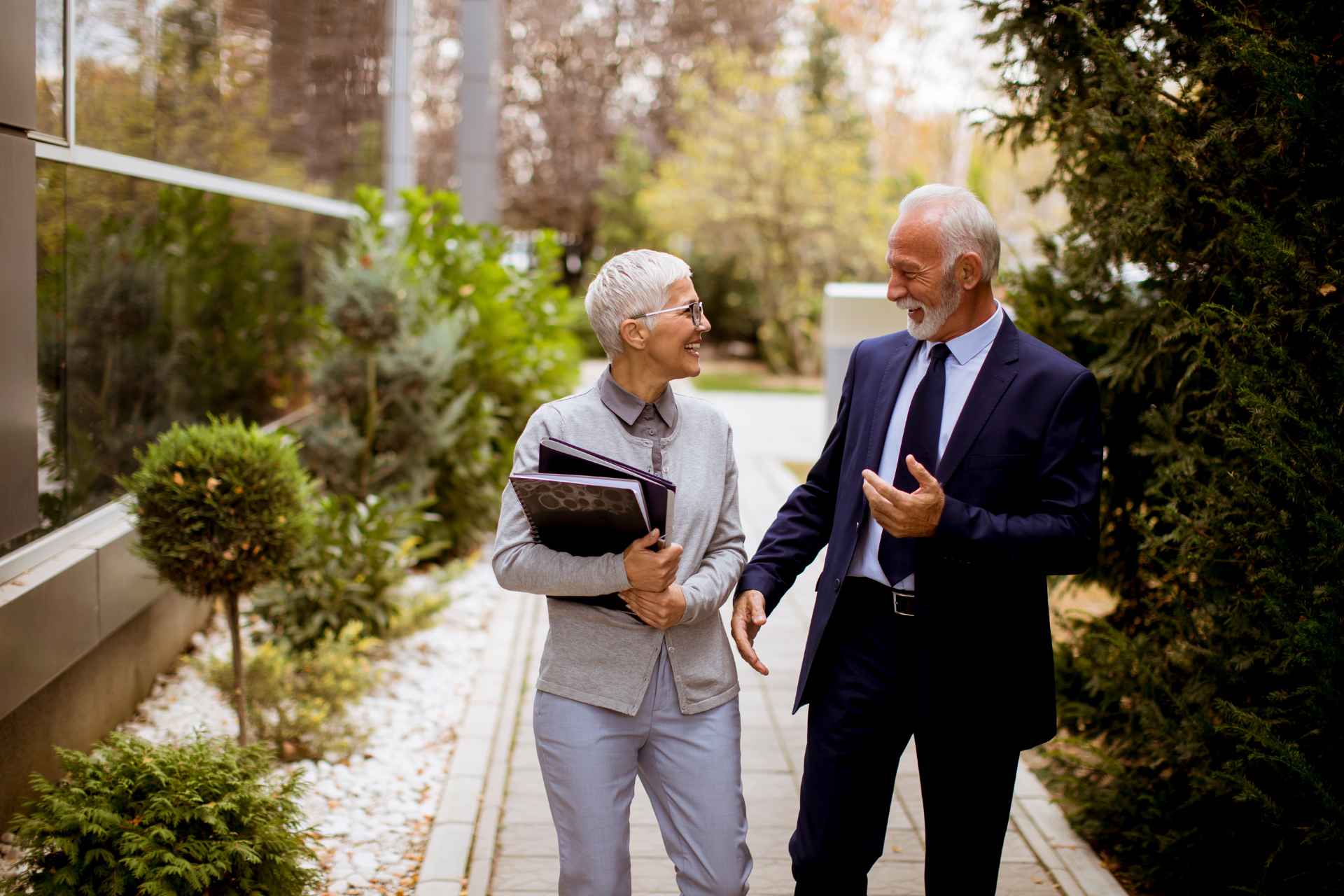 Two seniors in professional clothes are talking and laughing outside.