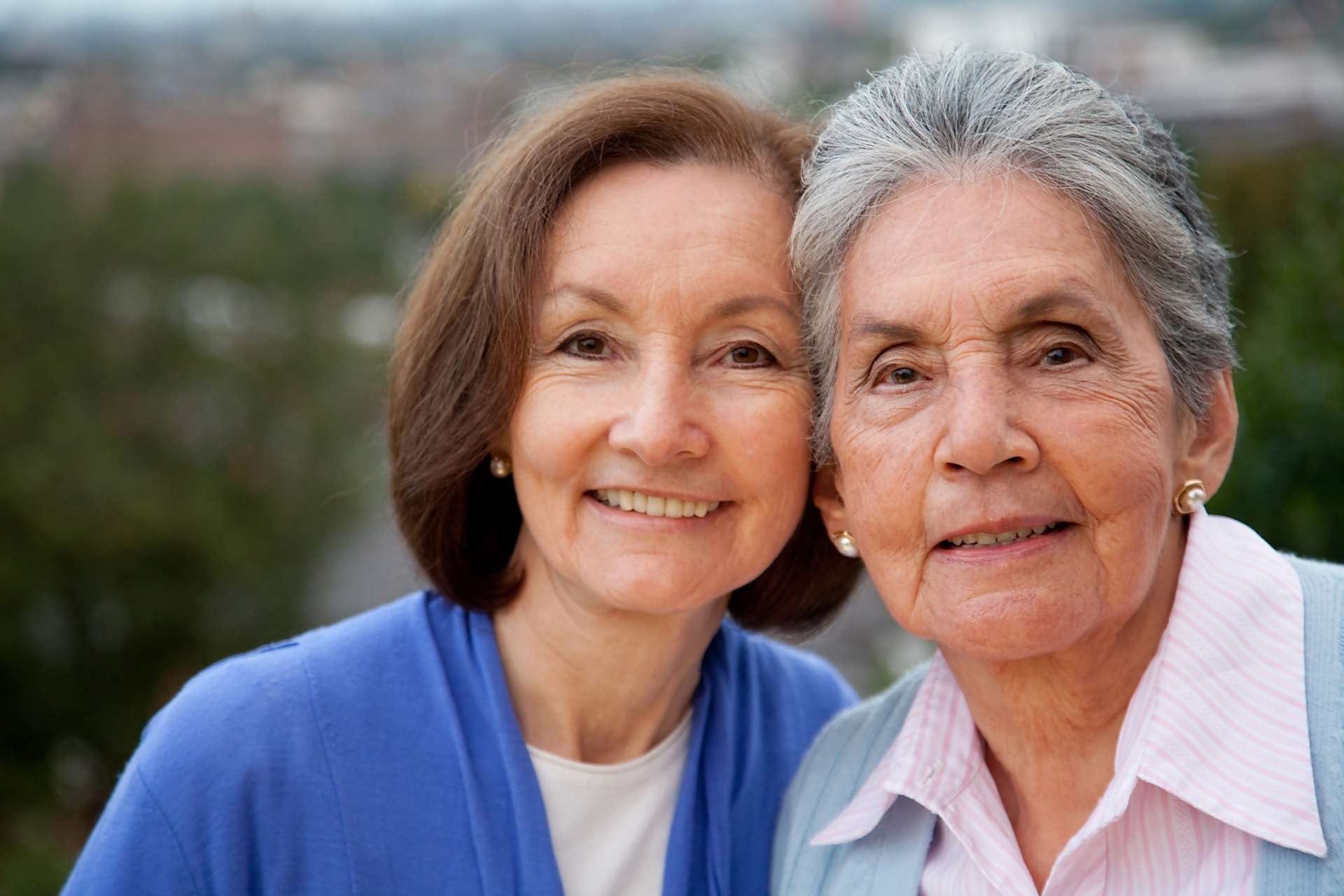 Two senior women smiling at the camera, leaning in towards each other.