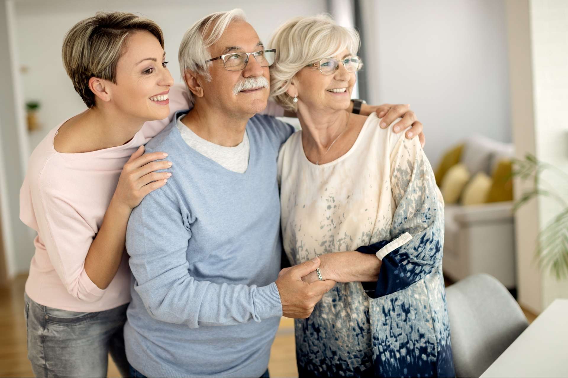 A senior couple and their daughter embracing and looking out a window.